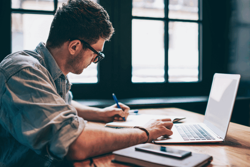 A young man implementing content marketing tips by creating a content calendar with a notebook and a laptop.
