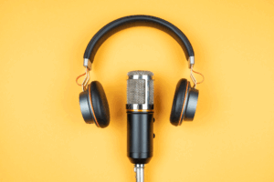 A black podcast microphone with a pair of headphones sitting over it so that the ear pieces sit either side of the microphone. This is on top of a vibrant yellow background.