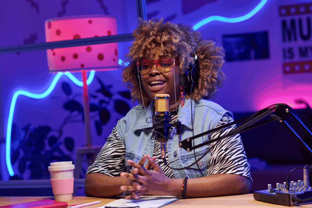 A trendy young woman recording a podcast with a smile on her face, a notebook in front of her, and a coffee to the side. The room is very modern, with blue and pink LED lights.
